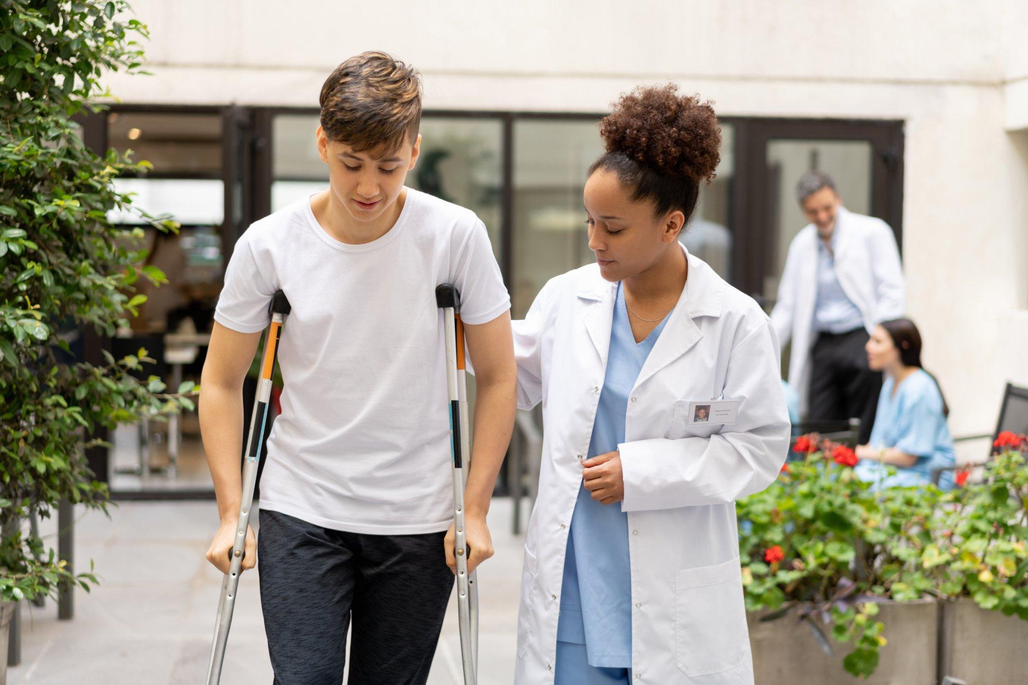 Friendly black female doctor assisting male teenager using crutches crossing the clinics patio