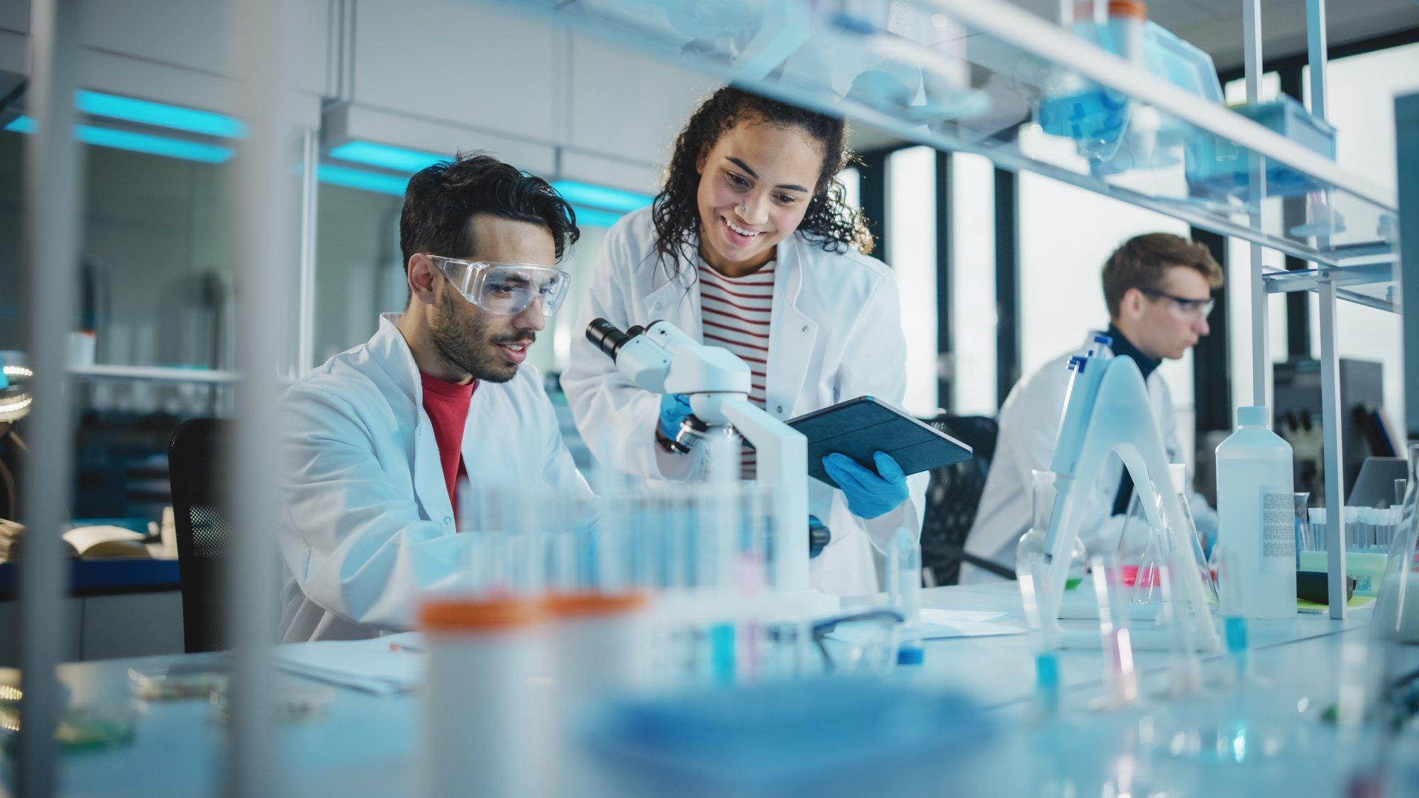 Modern Medical Research Laboratory Portrait of Latin and Black Young Scientists Using Microscope, Digital Tablet, Doing Sample Analysis, Talking. Diverse Team of Specialists work in Advanced Lab