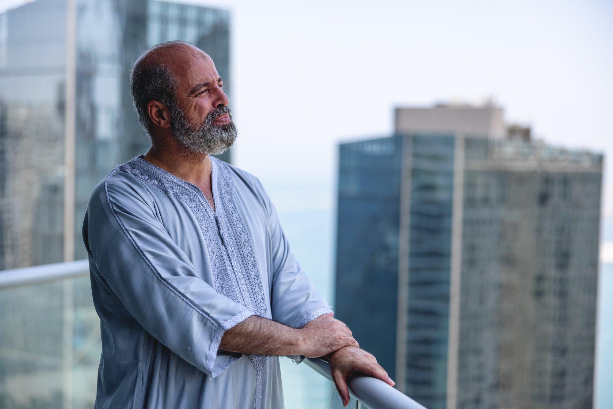 Portrait Of Older Middle Eastern Male On A Balcony Holding The Fence, Large Buildings In The Background